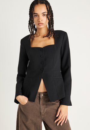 Zwarte getailleerde blazer met een vierkante halslijn, vijf knopen, dubbele voorkwasten en een slim-fit ontwerp. Draagt over een bruine broek.