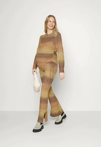 Mulher com um suéter de malha às riscas castanho e amarelo e calças largas, segurando uma pequena bolsa branca, a caminhar e a sorrir contra um fundo simples.