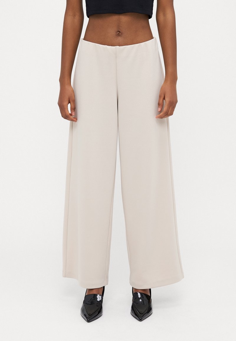Personne portant un pantalon beige à jambes larges, un haut court noir et des chaussures noires brillantes à bouts pointus, sur fond blanc uni.