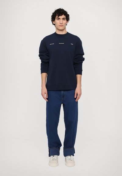 Námořnická modrá mikina s kulatým výstřihem, na přední straně s bílým textem. Kombinována s volnými modrými džíny a světle barevnými teniskami.