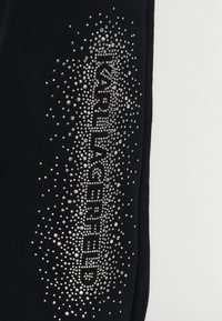 Μαύρα φόρμες με gradient από ασημένια ρουμπίνια, που γράφουν "Karl Lagerfeld", τοποθετημένα σε ένα λαμπερό, σημειακό σχέδιο κατά μήκος μίας πλευράς.