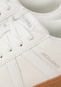 White leather sneakers with textured stitching, flat white laces, subtle branding, and a tan rubber sole. Simple design with three stripes.