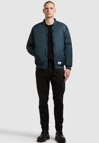 Man står och bär en teal bomberjacka, svart tröja, svarta byxor och svarta stövlar mot en enkel ljusgrå bakgrund.