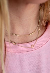 Zelta slāņa kaklarotas ar smalkām ķēdēm, horizontālu stieni un apaļām akcentiem, valkātas pretī rozā ribotā džemperī.