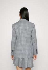 Blazer a cuadros con un corte ajustado, que presenta una solapa ancha, hombros estructurados y detalles de botones en las mangas. Se muestra vista trasera.