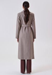 Lungo cappotto beige con cintura, dotato di colletto, maniche lunghe e taglio dritto. Texture liscia, la vista posteriore mostra dettagli delle cuciture.