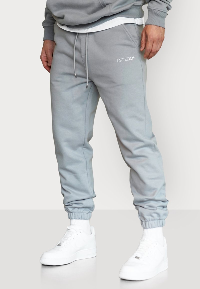 Mann trägt hellgraue Jogginghosen mit elastischen Bündchen und weißen Turnschuhen, steht vor einem schlichten Hintergrund.