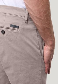 Beige katoenen broek met een smalle tailleband, een enkele achterzak met knoopsluiting en een zwart stoffen label met "pierre cardin" branding.