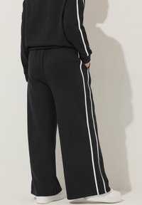Person med svarta vida byxor med vita sidremsor och matchande svart topp, stående med händerna i fickorna och vita sneakers.