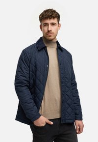 Marineblaue gesteppte Jacke mit einem Baumwollkragen, die mit Druckknöpfen geschlossen wird. Sie wird über einem beigen Rollkragenpullover und dunklen Jeans getragen, wobei die Taschen sichtbar sind.