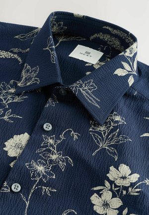 Chemise florale marine avec un tissu texturé, motif floral beige clair, deux boutons et un col classique avec une étiquette de marque à l'intérieur.
