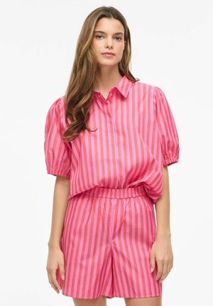 Nő hosszú barna hajjal, rózsaszín és piros, függőlegesen csíkos rövid ujjú ingben és hozzáillő, rugalmas derékpántos rövidnadrágban, fehér háttér előtt áll.