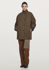 Brown padded coat with high collar, button closure, and side pockets. Paired with brown suede pants featuring a textured patch. Black footwear.