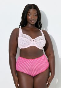 Pewna siebie kobieta plus size w różowym koronkowym biustonoszu i wzorzystych figach z wysokim stanem, pozująca na białym tle.