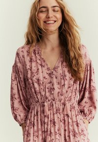 Lyserød blomsterkjole med V-udskæring, samlet bodice og lange klokkeærmer. Har frynse-detaljer og knapdetaljer. Blødt, tekstureret stof.