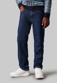 Jeans di cotone blu scuro con cinque tasche, cuciture a contrasto e design a gamba dritta, abbinati a sneakers bianche.