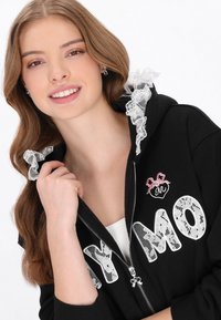 Sudadera negra con cremallera y capucha, con letras superpuestas de encaje blanco que dicen "MY MO", un pequeño detalle bordado de corazón y acentos de encaje en la capucha.