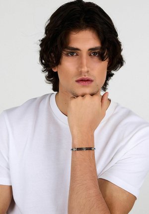 Bracciale in pelle nera con dettagli in metallo color argento, caratterizzato da una superficie liscia e accenti metallici. Indossato su un polso con una maglietta bianca.