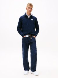 Pulôver de fleece da marinha com gola de fecho e logo no peito, emparelhado com calças cargo azul-escuras e ténis brancos.