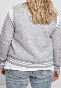Sweatshirt gris en tissu doux, avec des accents rayés blancs sur les poignets et l'ourlet, superposé sur une chemise blanche, associé à un jean en denim.