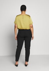 Olivgrön kortärmad blus med ett nyckelhål i ryggen, kombinerad med åtsittande svarta byxor och svarta sandalett med öppen tå. Slät tygtextur.