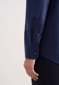 Granatowa koszula z długim rękawem z ostro zakończonym mankietem zapinanym na dwa dopasowane guziki oraz gładką fakturą tkaniny. Ręka spoczywa w pobliżu dolnego brzegu koszuli.