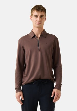 Mann trägt ein braunes Langarm-Poloshirt mit Reißverschluss und dunkle Hose, steht vor einem einfarbigen hellgrauen Hintergrund.