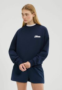 Mujer con cabello largo y rubio, vistiendo sudadera de Ellesse color azul marino y pantalones cortos a juego, mirando hacia la izquierda con las manos entrelazadas delante.