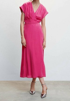 Femme portant une robe midi portefeuille rose vif à manches courtes et des escarpins argentés à bout pointu avec bride à la cheville, debout devant un fond uni.