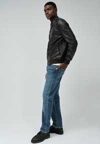 Bruine leren bomberjack, blauwe spijkerbroek, zwarte schoenen. Het jack heeft een ritszak, ribbelmanchetten en een kraagdetail.