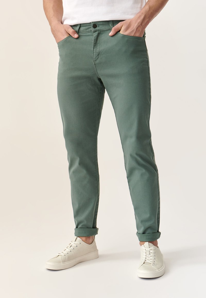 Pantalones de mezclilla verdes con un corte ajustado, diseño de cinco bolsillos y puños enrollados, combinados con zapatillas blancas con cordones.