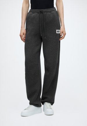 Personne portant un pantalon de survêtement gris foncé avec une taille à cordon et des baskets blanches sur un fond blanc uni.