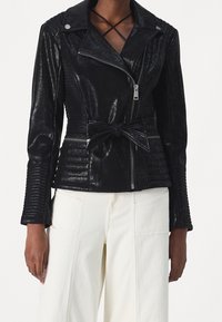 Giacca biker in finta pelle nera con vita regolabile, texture a coste sulle maniche, zip asimmetrica e dettagli in metallo sui bottoni.
