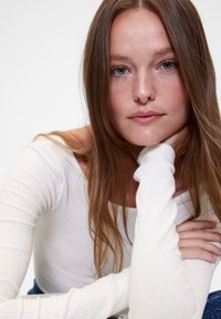 Biały prążkowany top z długim rękawem i dopasowanymi rękawami, z okrągłym dekoltem. Modelka ma długie, brązowe włosy i opiera brodę na dłoni.