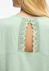Jasnozielony top z wycięciem na plecach ozdobionym misterną koronką. Materiał ma teksturowane wykończenie i luźny krój.