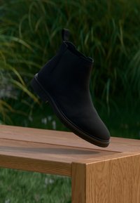 Melna potītes zābaki ar elastīgām sānu panelēm, gludu tekstūru, apaļu purngalu un plānu gumijas zoli, novietoti uz koka virsmas.