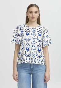 Top a maniche corte in bianco con motivo ikat blu e blu navy. Vestibilità ampia, scollatura rotonda e tessuto testurizzato. Indossato con jeans azzurri.