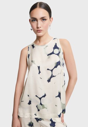 Mladá žena s uhladenými tmavými vlasmi, oblečená v tielkovej blúzke a zodpovedajúcich nohaviciach s bielym a námornícky modrým abstraktným kvetinovým vzorom.