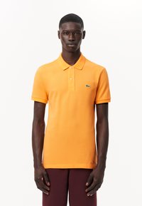 Orange pikétröja med korta ärmar och krage, med en grön broderad logotyp på vänster bröst, i mjukt bomullstyg.