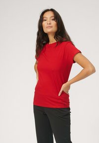 Rød t-shirt med korte ærmer lavet af blødt stof, med rund hals og en tætsiddende pasform. Båret med sorte bukser, vist fra siden.