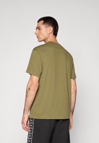 T-shirt verde-oliva com mangas curtas, gola redonda e corte descontraído, feita de tecido de algodão macio. Tatuagens visíveis no braço direito.