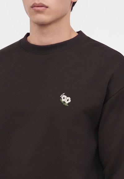 Jeune personne portant un sweat-shirt noir à col rond avec une petite broderie de fleur blanche sur la poitrine gauche.