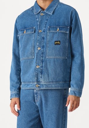Homme portant une veste en jean bleue et un jean assorti, avec des poches à rabats et des boutons à l'avant de la veste.