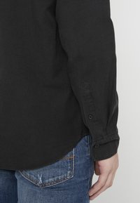 Czarna koszula z długim rękawem z mankietem na guziki, wykonana z teksturowanego materiału, zestawiona z niebieskimi dżinsami z postrzępioną kieszenią.