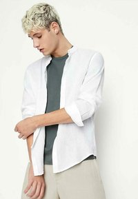 Camicia bianca in lino con una vestibilità morbida e colletto a fascia, indossata sopra una maglietta verde scuro. Il modello indossa pantaloni beige chiaro.