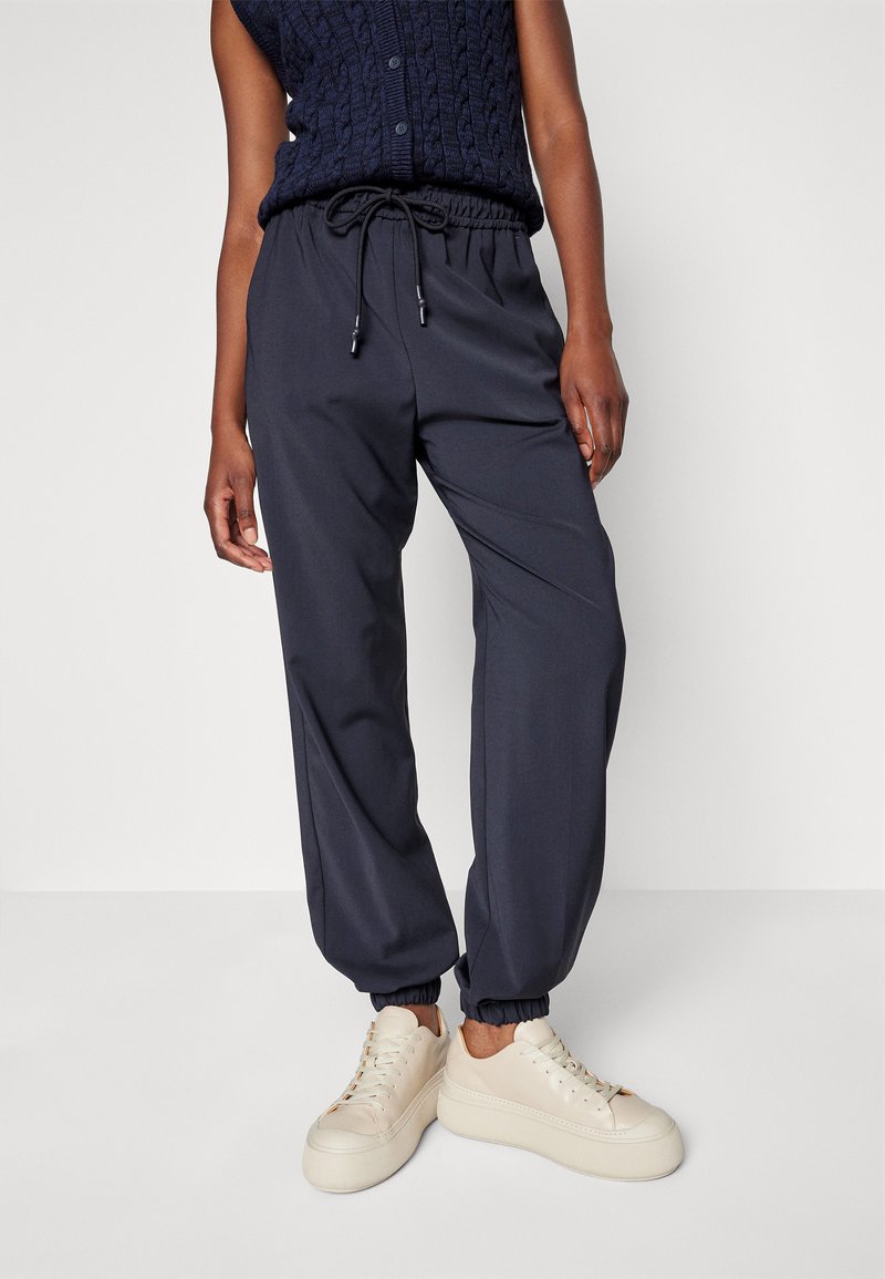 Granatowe spodnie dresowe z elastycznym paskiem w talii i regulowanym sznurkiem. Posiadają ściągacze na nogawkach oraz gładki, lekki materiał. Noszone z beżowymi trampkami.