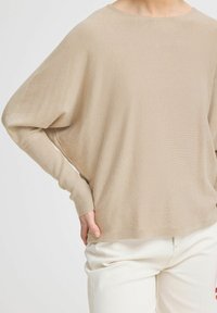Pull en tricot lâche beige avec un col large et des épaules tombantes, doté de poignets côtelés et d'une finition douce et texturée. Associé à un jean clair.