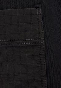 Zbliżenie czarnej kieszeni z tkaniny z przeszywanym wzorem w kratkę na teksturowanym czarnym tle tkaniny.