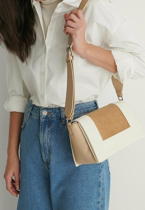 Vrouw draagt een wit shirt en blauwe spijkerbroek en houdt een beige en witte schoudertas met brede band over de schouder.