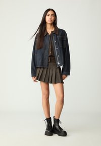 Giacca di jeans con colletto in velluto a coste marrone, abbinata a una camicia marrone con bottoni e una gonna plissettata scura. Stivali neri con lacci.